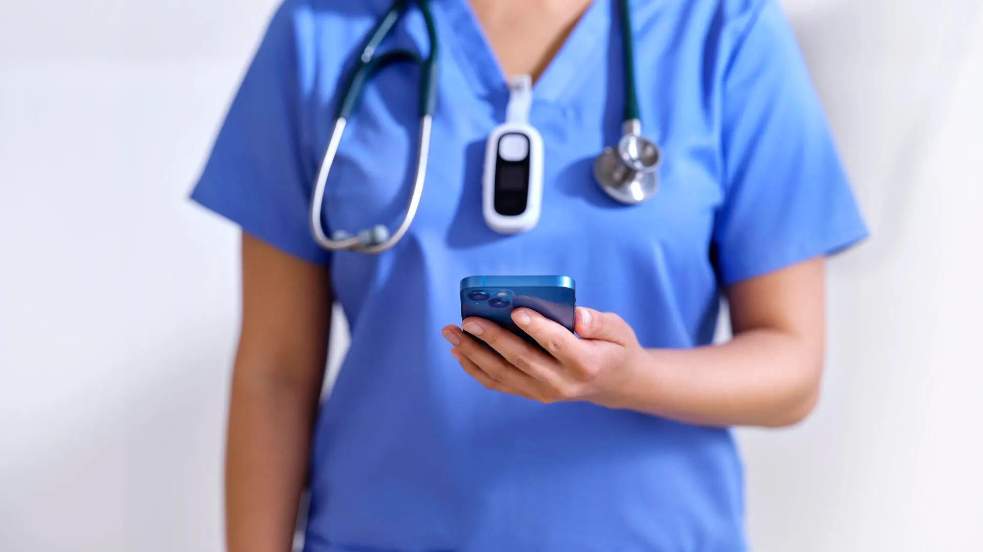 A healthcare worker in scrubs and wearing a tracking device looks at their mobile phone